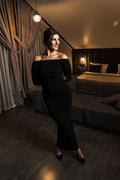 Κομψή γυναίκα με μαύρο φόρεμα που ποζάρει σε διαμέρισμα. Έννοια λήψης μόδας - Φωτογραφία, εικόνα