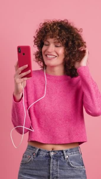 スマートフォンを使ってカーリーヘアでピンクの服を着た陽気な若い女性と,パステルカラーの背景に対してヘッドフォンを通して音楽を聴く - 映像、動画