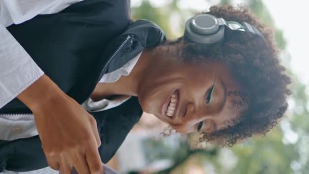 Alegre dama escuchando música por auriculares inalámbricos que sostienen el teléfono móvil de cerca. Mujer afroamericana feliz usando aplicación telefónica en la calle de la ciudad orientada vertical. Chica positiva disfrutar de la canción favorita - Metraje, vídeo