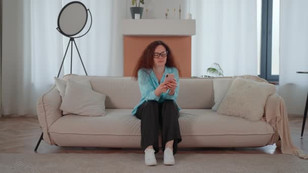 nainen punainen kihara tukka tilalla älypuhelin chattailuun ystävän kanssa verkossa viestintä istuu sohvalla asunnossa - Materiaali, video