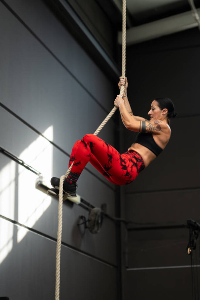 Vertical photo of a strong mature woman with tattooed arms climbing a rope in a cross training gym - Photo, Image