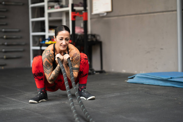 Strong woman squatting using a battle rope to work out in a gym - Photo, Image