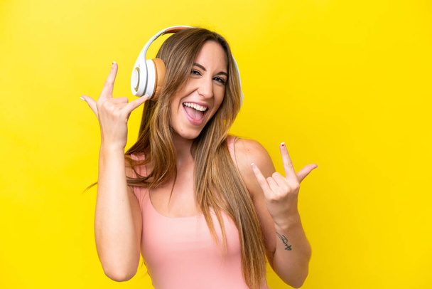 Mujer joven caucásica aislada sobre fondo amarillo escuchando música haciendo gestos de rock - Foto, imagen