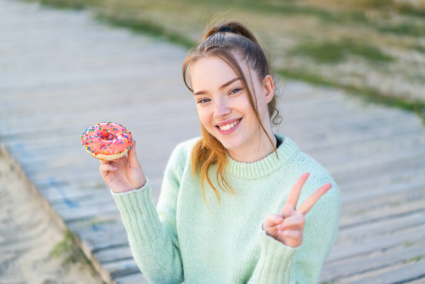 Genç ve güzel bir kız elinde donut, gülümsüyor ve zafer işareti gösteriyor. - Fotoğraf, Görsel