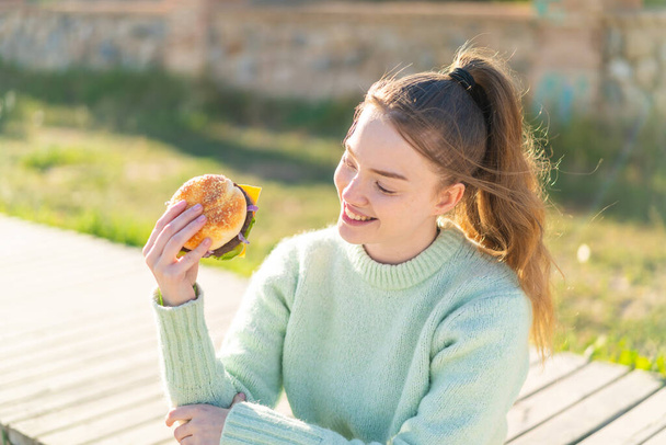 Νεαρό όμορφο κορίτσι κρατώντας ένα burger σε εξωτερικούς χώρους με ευτυχισμένη έκφραση - Φωτογραφία, εικόνα