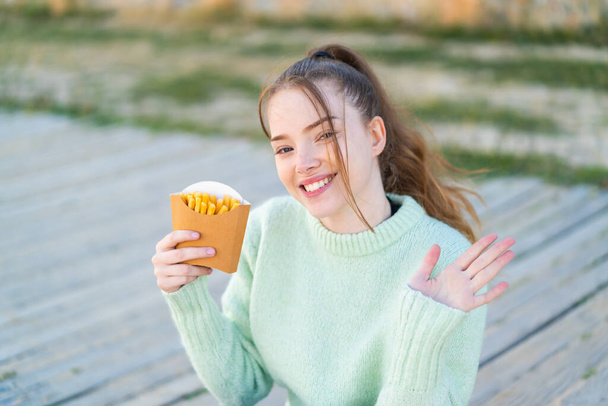 Νεαρό όμορφο κορίτσι κρατώντας τηγανητά τσιπς σε εξωτερικούς χώρους χαιρετισμό με το χέρι με ευτυχισμένη έκφραση - Φωτογραφία, εικόνα