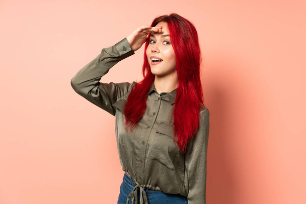 Teenager red hair girl isolated on pink background doing surprise gesture while looking to the side - Photo, Image