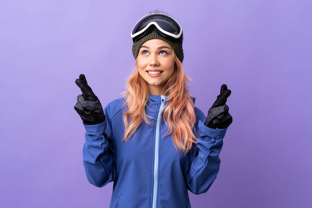 Skier adolescente avec des lunettes de snowboard sur fond violet isolé avec les doigts croisés et souhaitant le meilleur - Photo, image