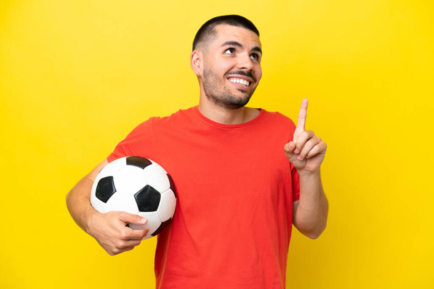 Junger kaukasischer Mann spielt Fußball isoliert auf gelbem Hintergrund und weist auf eine großartige Idee hin - Foto, Bild