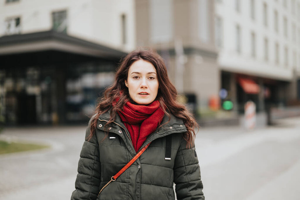 Young woman in warm jacket and red scarf outdoors, city life concept, focused and serious expression. - Photo, Image