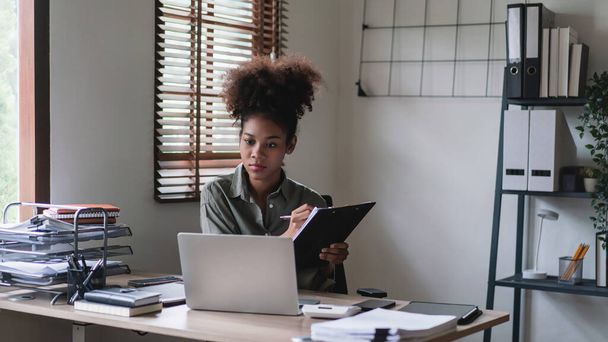 Femme d'affaires afro-américaine lisant des données commerciales financières sur ordinateur portable et écrivant une note dans le document. - Photo, image