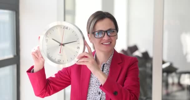 Krásná usměvavá žena v kanceláři drží stylové nástěnné hodiny. Přesčasová lhůta a obchodní žena - Záběry, video