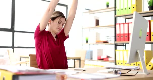 La mujer estira los brazos hacia arriba en el lugar de trabajo en la oficina. Gerente toma un descanso haciendo ejercicios simples sentado en el lugar de trabajo - Imágenes, Vídeo