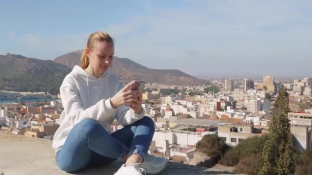 szczęśliwa dziewczyna turystyczna siedzi, podziwiając widok na miasto z góry, trzymając telefon w rękach, aparat zbliża się do dziewczyny. Wysokiej jakości materiał 4k - Materiał filmowy, wideo