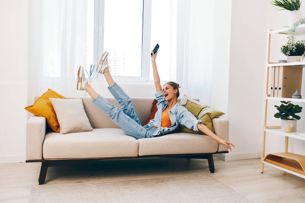 Glimlachende vrouw met een mobiele telefoon, genieten van online games en chatten tijdens het zitten op een gezellige bank in een stijlvolle woonkamer. - Foto, afbeelding