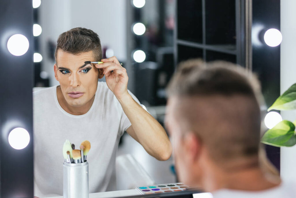 Reflexión en un espejo de un hombre gay moldeando sus cejas mientras hace maquillaje para un espectáculo - Foto, Imagen