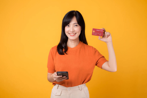 Excited Asian woman 30s, using smartphone and showing credit card on yellow background. Convenient online shopping - Photo, Image