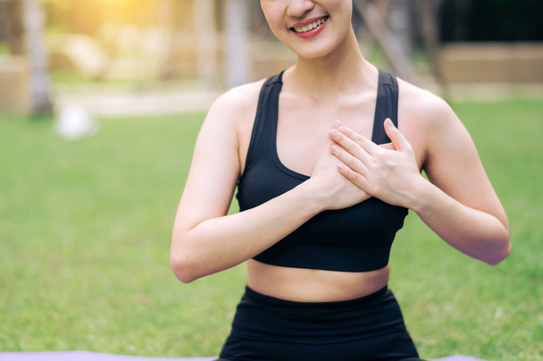 Retrato ajuste jovem asiático mulher 30 anos vestindo sportswear preto de mãos dadas no peito enquanto escuta relaxar música e sentado no tapete de ioga no parque público. Conceito de bem-estar de saúde. - Foto, Imagem
