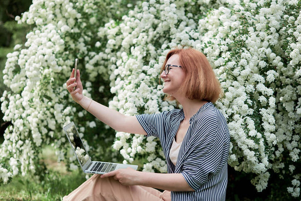 Ανεξάρτητος τρόπος ζωής, απομακρυσμένη εργασία σε πλήρη άνθιση μια νεαρή γυναίκα αιχμαλωτίζει τη στιγμή της ανάμεσα σε λευκά λουλούδια με φορητό υπολογιστή. - Φωτογραφία, εικόνα