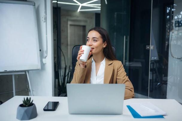 Νεαρή επιχειρηματίας που φοράει μπεζ κοστούμι πίνοντας καφέ στο γραφείο. Ευτυχισμένη χαμογελαστή γυναίκα επαγγελματίας εργασίας ετοιμαστείτε για παραγωγική ημέρα - Φωτογραφία, εικόνα