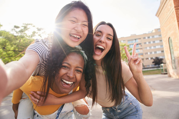 Tři mladí multiraciální přátelé, kteří spolu v létě fotí selfie portrét venku. Latinská žena dává svezení na zádech asijské ženy na ulici města. Nadšené dívky pózující pro legrační fotografii, objektiv světlice. - Fotografie, Obrázek