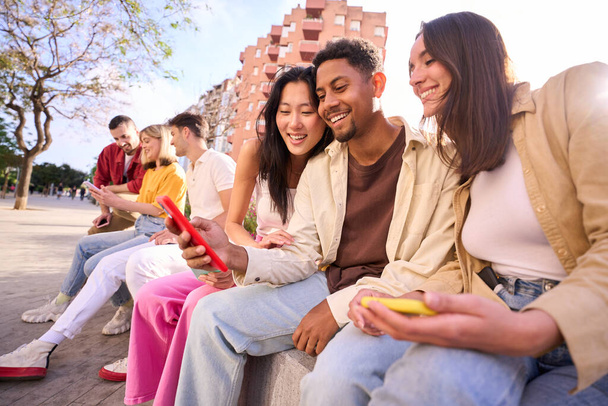 Junge Gruppe von Millennials, die sich draußen mit dem Handy vergnügen. Fröhliche Gemeinschaft von Studenten, die Social-Media-Inhalte auf dem Smartphone genießen. Multiethnische Freundesgruppe schaut mobil zu - Foto, Bild