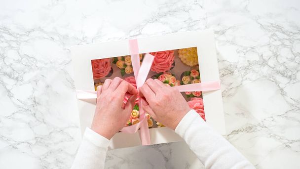 Acostado. Las manos de una mujer abren suavemente una caja de papel blanco, revelando un conjunto de cupcakes gourmet en el interior, cada uno artísticamente adornado con glaseado de crema de mantequilla, diseñado para imitar vibrantes rosas y tulipanes. - Foto, Imagen