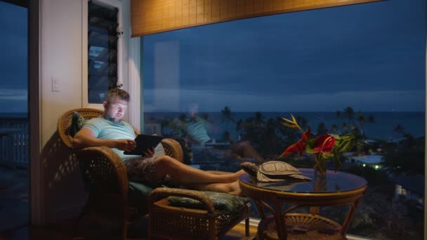 Guy erkundet das Reiseziel online. Männliche Suchergebnisse auf dem Tablet in einem Zimmer im hawaiianischen Inselstil. Mann sitzt am großen Wandfenster mit Meerblick und schaut nachts auf Tablet-Bildschirm mit WiFi - Filmmaterial, Video