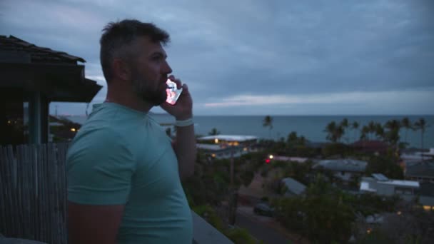 Ein bärtiger, gesunder Kerl steht an einem bewölkten Tag auf einem Balkon mit Blick auf die tropische Insel. Sportlich lächelnder Typ, der draußen mit dem Smartphone spricht. Geschäftsmann Freiberufler, der aus der Ferne arbeitet, beantwortet Anruf - Filmmaterial, Video