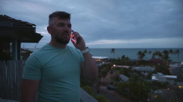 Bearded businessman free lancer working remotely answer call. Positive healthy guy standing on balcony with tropical island views on overcast day. Athletic smiling guy talking outside using smartphone - Footage, Video