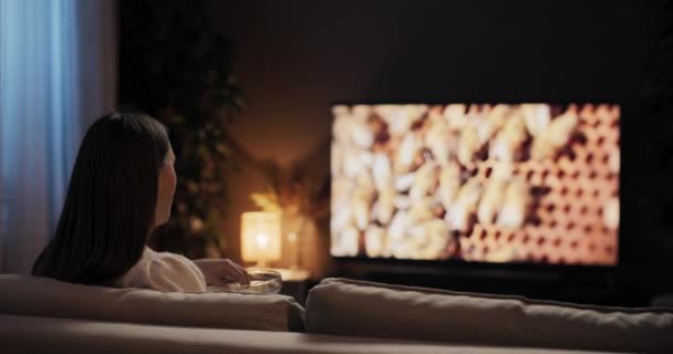 Attraktive College-Studentin, die eine Auszeit vom Studium für Prüfungen nimmt, sitzt im gemütlichen Wohnzimmer und schaut Lieblingsfernsehsendung beim Naschen von Popcorn. - Filmmaterial, Video