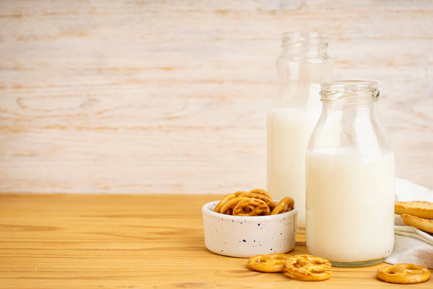 Melk in glas en kan met koekje op houten tafel en lichte ondergrond. Close-up. - Foto, afbeelding