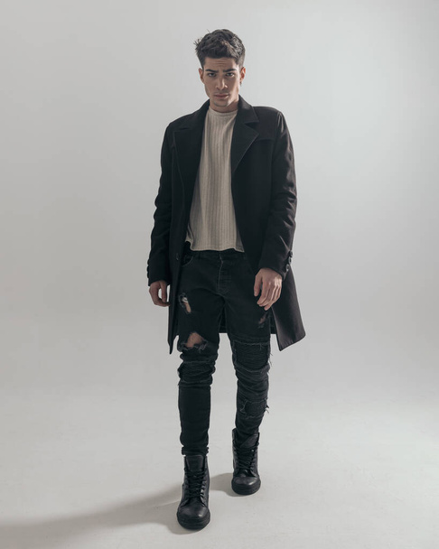 selbstbewusster und sexy junger Mann in schwarzem Mantel und Jeans, der sich auf grauem Hintergrund vorwärts bewegt - Foto, Bild
