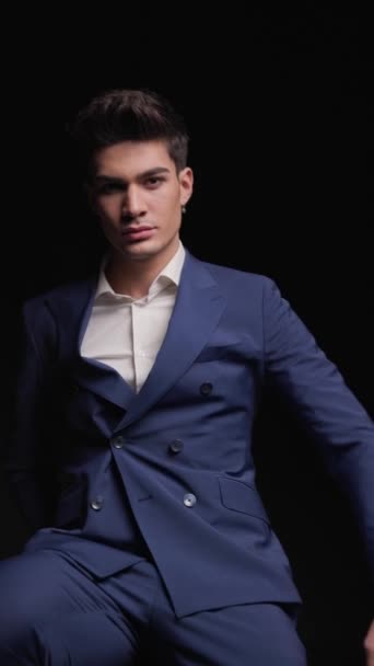 sexy young man in dobule breasted blue suit leaning forward and touching his chin while sitting on black background - Footage, Video
