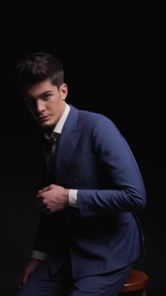 confident elegant man in blue suit sitting, holding elbow on knee, touching chin and being thoughtful while looking away on dark background - Footage, Video