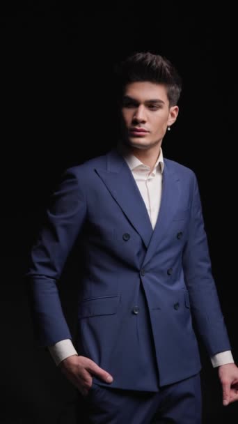 relaxed fashion male model in double breasted blue suit turning away from camera and walking into dark background - Footage, Video