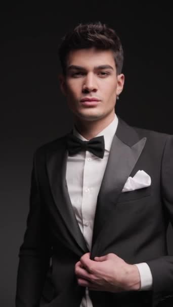 elegant man buttoning his black tuxedo while standing with hands in pockets on dark background with lightning effects - Footage, Video