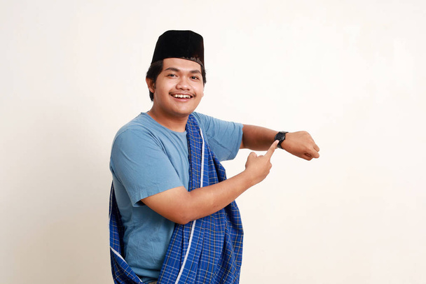 feliz asiático musulmán chico de pie mientras apuntando y mostrando su reloj de pulsera - Foto, Imagen