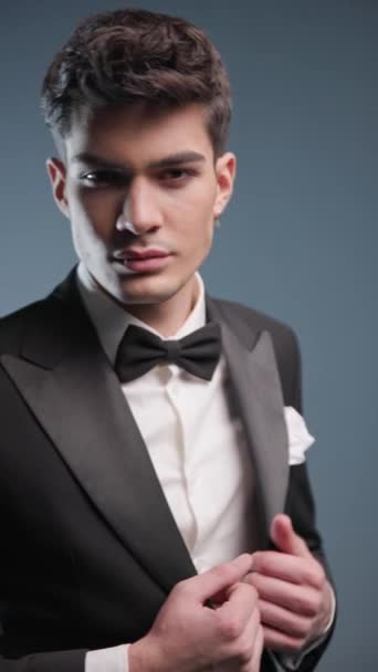 handsome elegant groom wearing black tuxedo, holding hands in pockets, looking down and fixing tux in front of grey background with flash lights on - Footage, Video