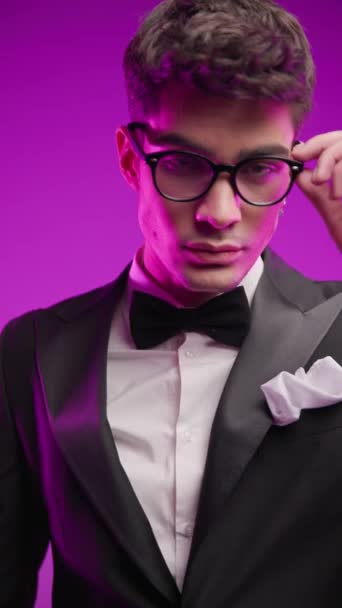 elegant businessman in black tuxedo adjusting eyeglasses, holding hand in pocket and looking to side while fixing black tuxedo in front of colorful background - Footage, Video
