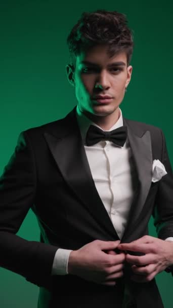 elegant businessman unbuttoning black tuxedo, looking forward and being confident in front of colorful background - Footage, Video