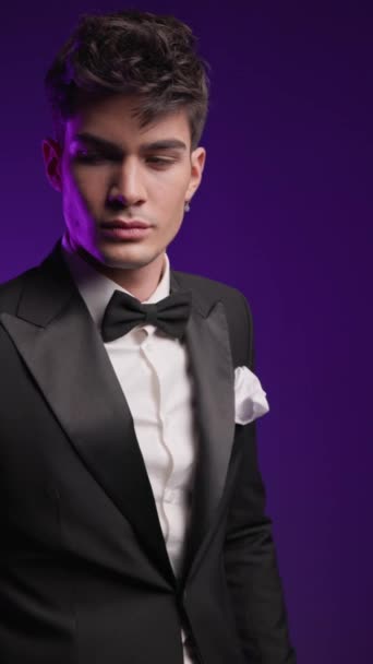 attractive young groom posing and adjusting black tuxedo, looking to side, holding hand in pocket in front and moving in front of colorful background - Footage, Video