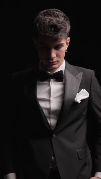 handsome elegant man looking down, holding hands in pockets, looking forward while wearing black tuxedo in front of dark background - Footage, Video