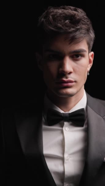 close up of handsome young man wearing black tux and bowtie standing with hands in pockets on black background and looking down - Footage, Video