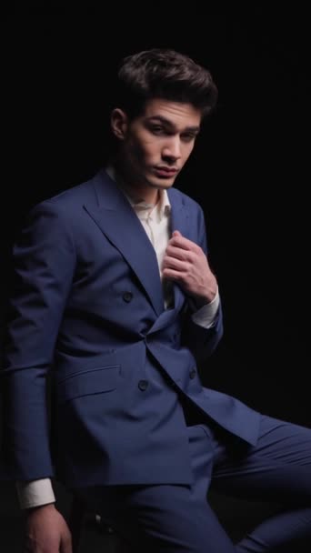sexy businessman adjusting blue suit, being confident, smiling and looking to side while sitting and posing in front of dark background - Footage, Video