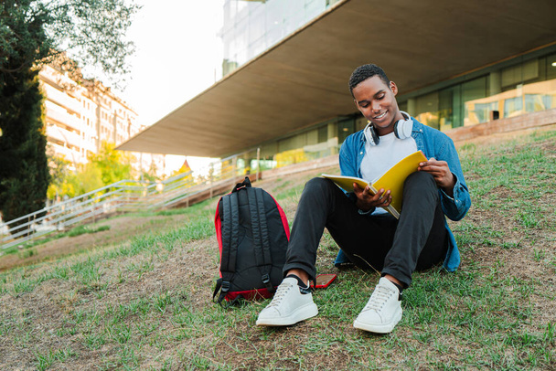 Afroameričan chytrý teenager čte knihu, která sedí na univerzitním kampusu. Fešák koncentrovaný mladý muž s brýlemi studuje a učí se její poznámky z lekce na středoškolské zkoušky - Fotografie, Obrázek