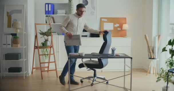 Nuori mies liike-elämän ammattilainen pitää kannettavan tietokoneen ja älypuhelimen pöydällä saapuessaan toimistoon - Materiaali, video