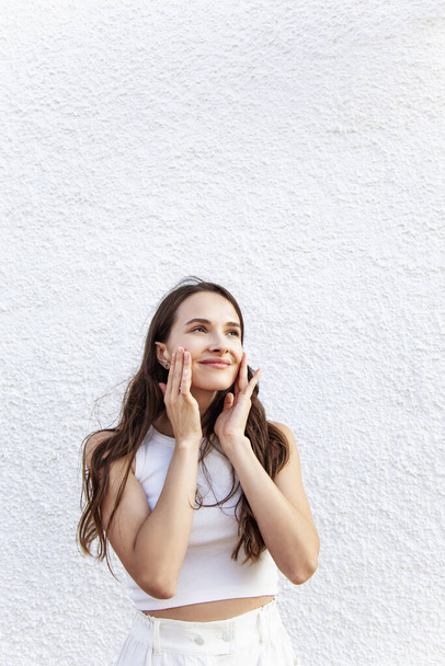 beautiful young brunette with hands on face in white top looking to side and smiling on white wall background - 写真・画像
