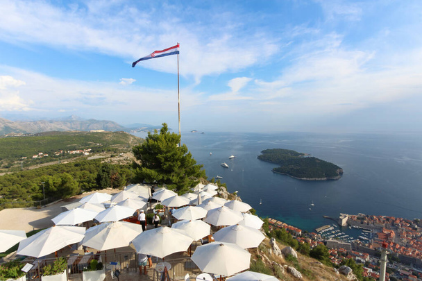 Experimente o encantador Dubrovnik, onde as majestosas paisagens urbanas encontram tranquilos litorais e pitorescas montanhas. A orgulhosa bandeira croata acena graciosamente, oferecendo um vislumbre da icónica localização de Game of Thrones. Experimente o fascínio de Dubrovni - Foto, Imagem