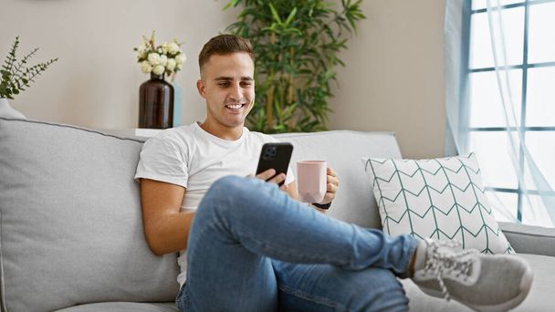 スマートフォンを使用して微笑む若いヒスパニック男性,コーヒーマグで自宅でソファーでリラックス. - 写真・画像
